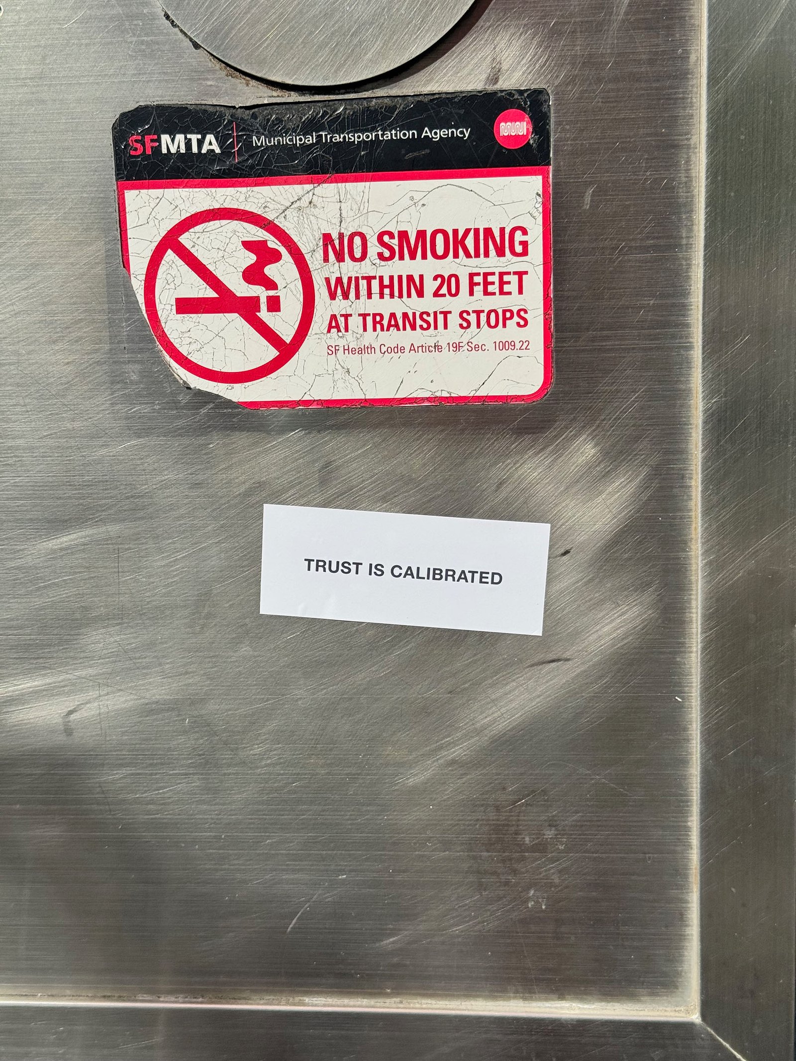 Control Text field test sticker reading “Trust Is Calibrated” placed on a metal surface beneath a municipal no smoking sign in San Francisco.